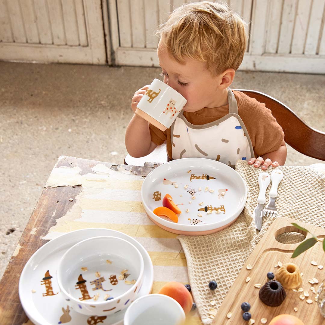 Een peuter drinkt uit de lässig porseleinen mok van de serviesset met dierenpatroon aan een houten tafel.