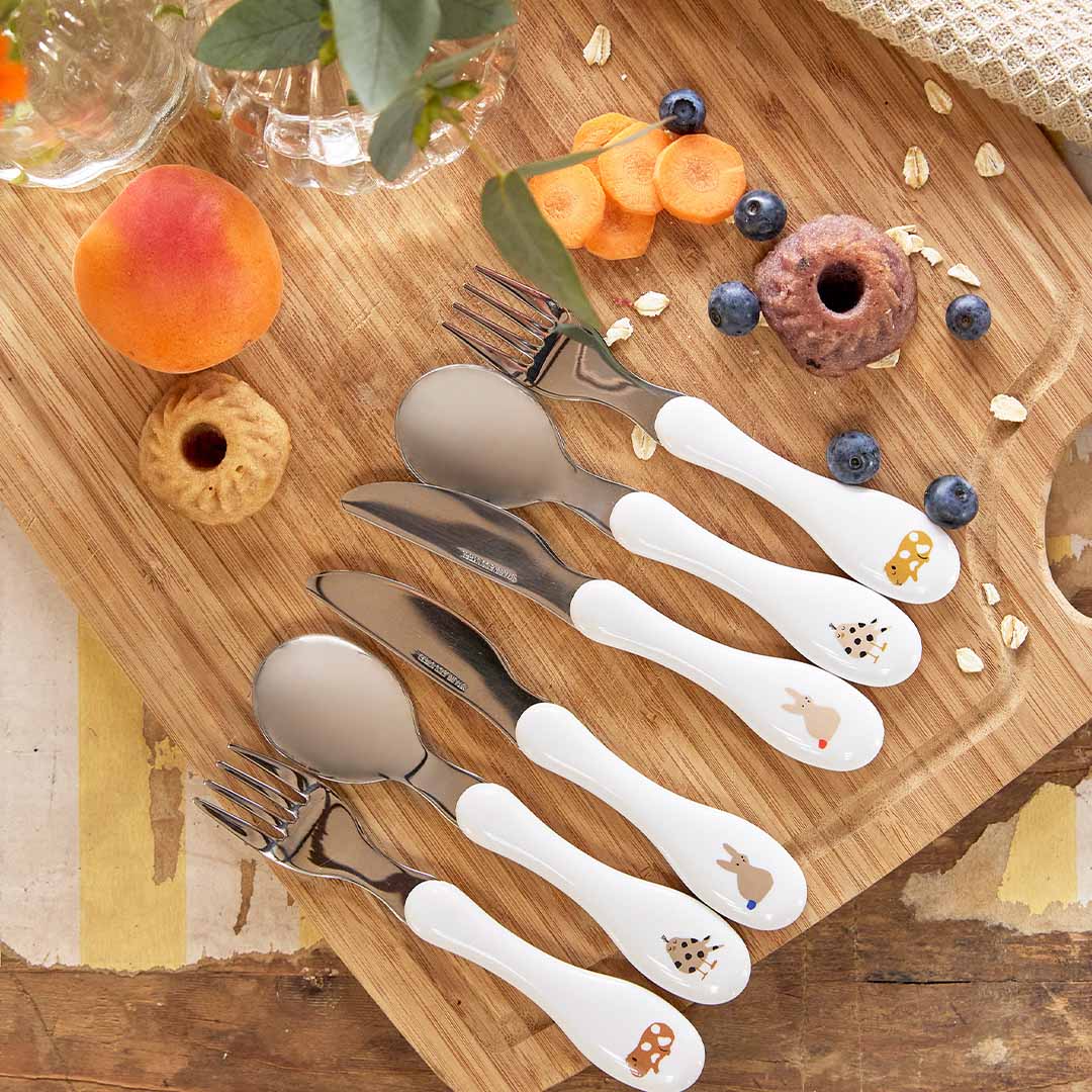 Twee sets lässig kinderbestek met dierenpatronen op een houten plank met fruit en snacks.