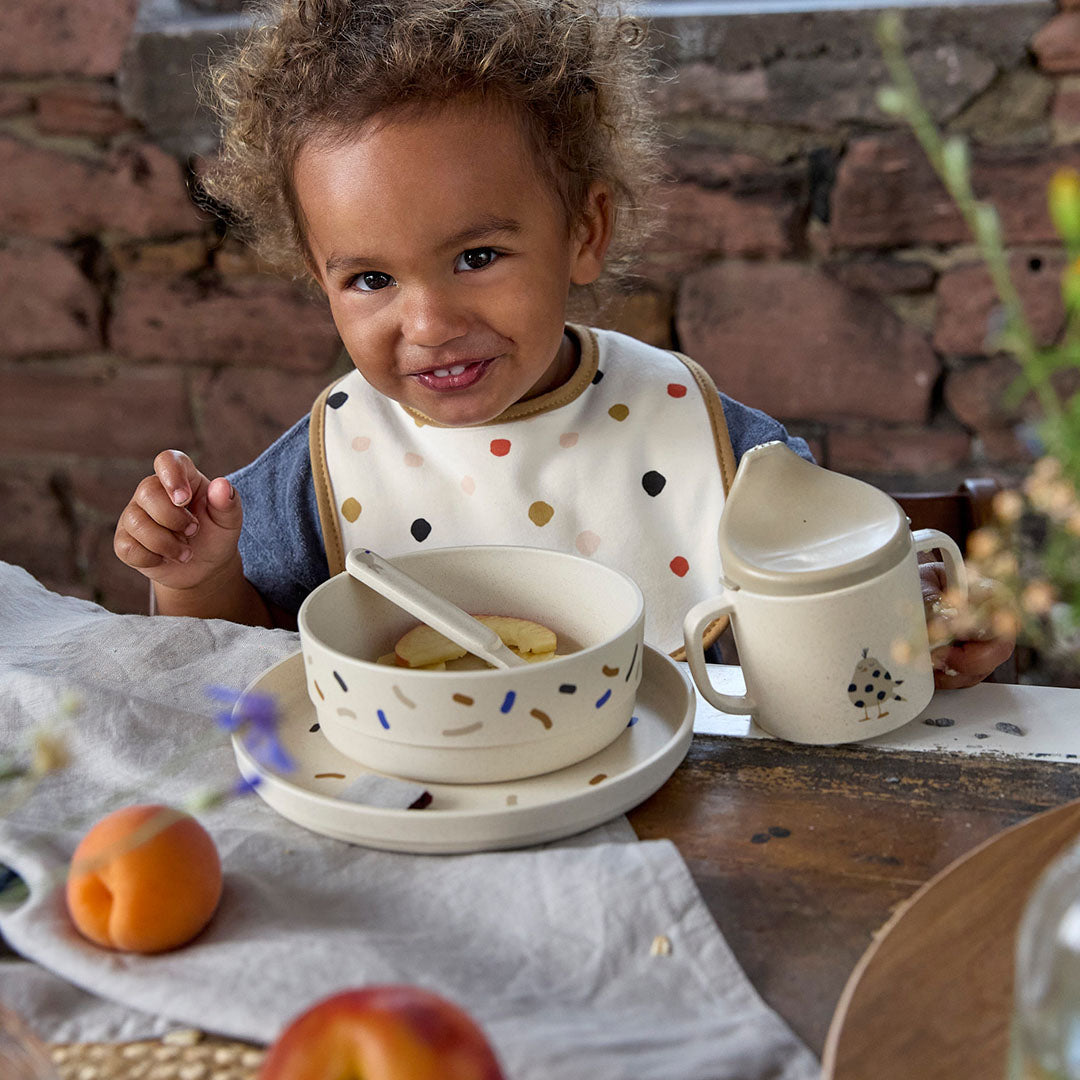 Een lachende peuter zit aan tafel met een lässig babyserviesset met spikkels en een lieveheersbeestje op de drinkbeker.