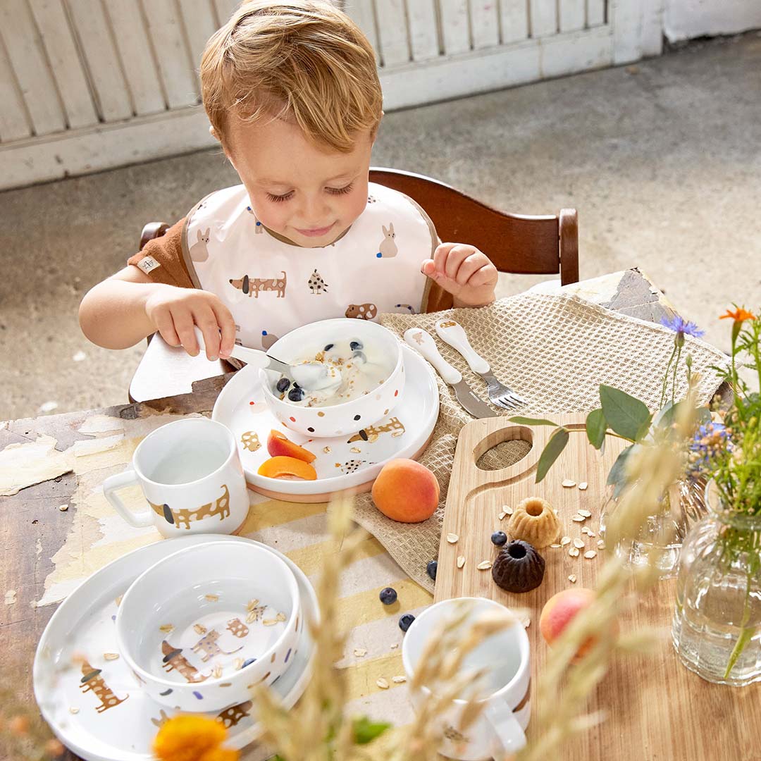 Een peuter eet met een lepel uit de lässig porseleinen kom van de serviesset met dierenpatroon aan een houten tafel.