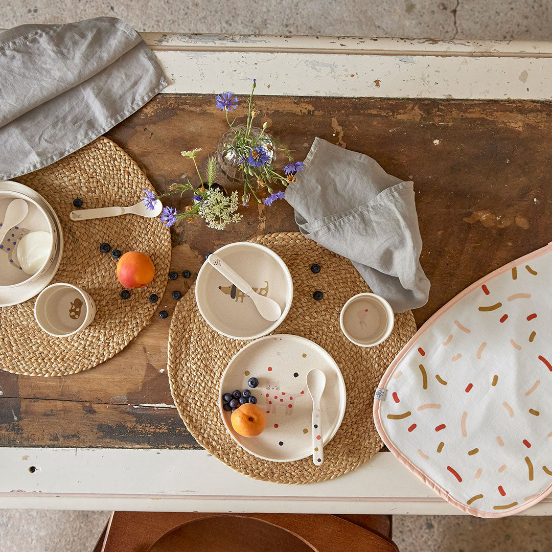Twee lässig babyserviessets met dieren en stippen op een houten tafel, omringd door fruit en bloemen.