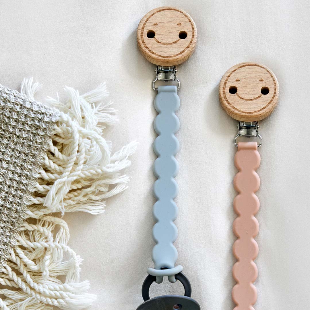 Twee Lässig speenkoorden met houten smiley clips en siliconen banden in blauw en roze, liggend op een deken.