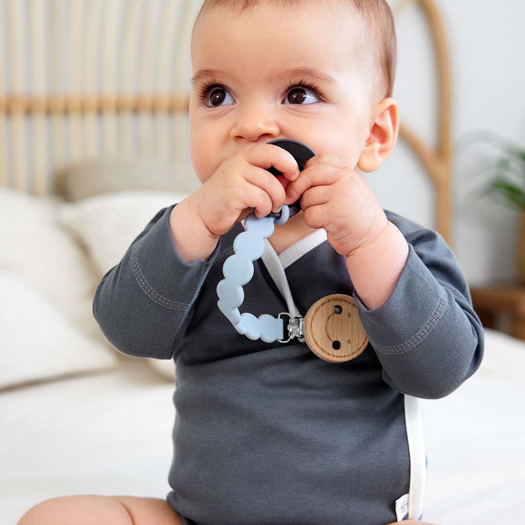 Een baby met een donkergrijze romper draagt het lässig speenkoord met een donkergrijze speen.