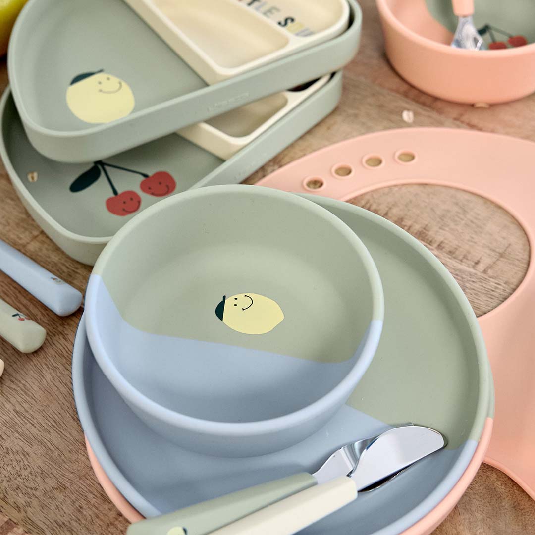 Een collectie Lässig voedingsbenodigdheden voor baby's en peuters, waaronder borden, kommen, een slab en bestek met kersen- en citroenmotieven.