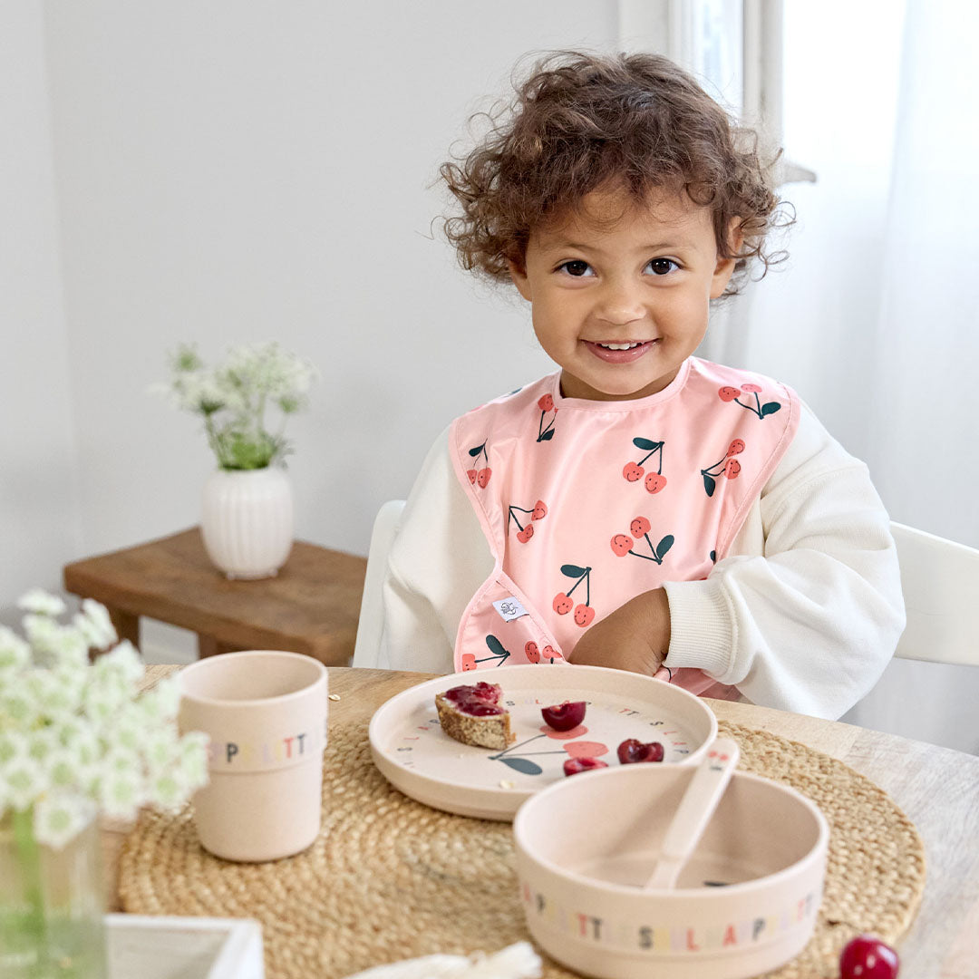 Een lachend kindje met krullen draagt een roze slab en zit aan tafel met de Lässig beige serviesset met kersenpatroon.