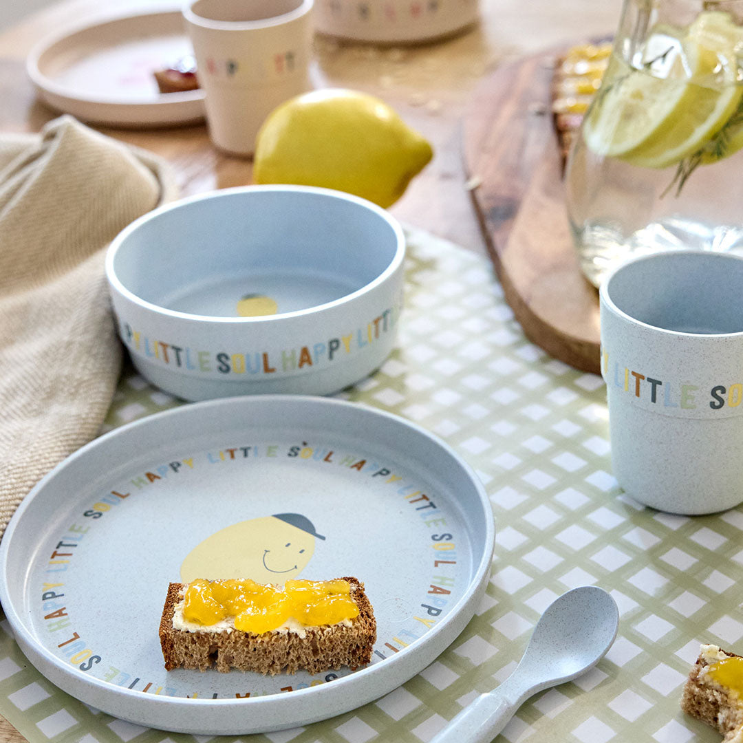 De Lässig lichtblauwe baby serviesset met citroenprint staat op een tafel met een boterham met jam en een glas water.