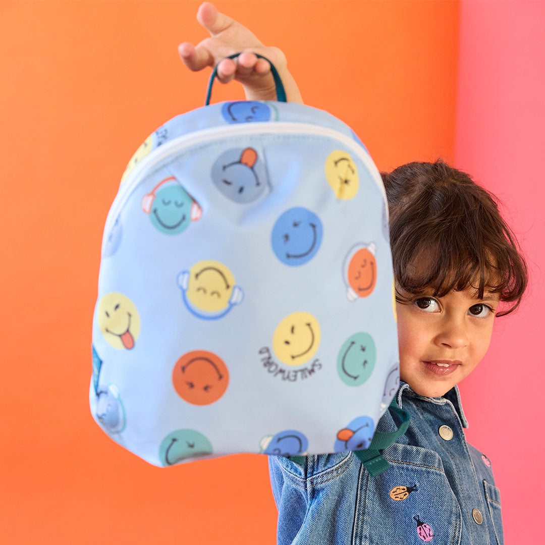 Een kind houdt de blauwe peuterrugzak met smileyworld-print schuin voor zich uit.