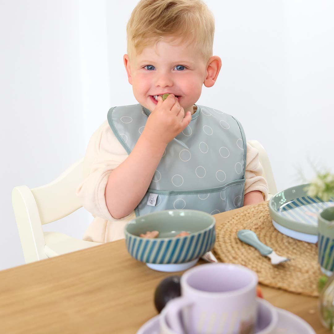 Een lachend kindje met een Lässig slabber eet aan tafel, met de porseleinen serviesset op de achtergrond.
