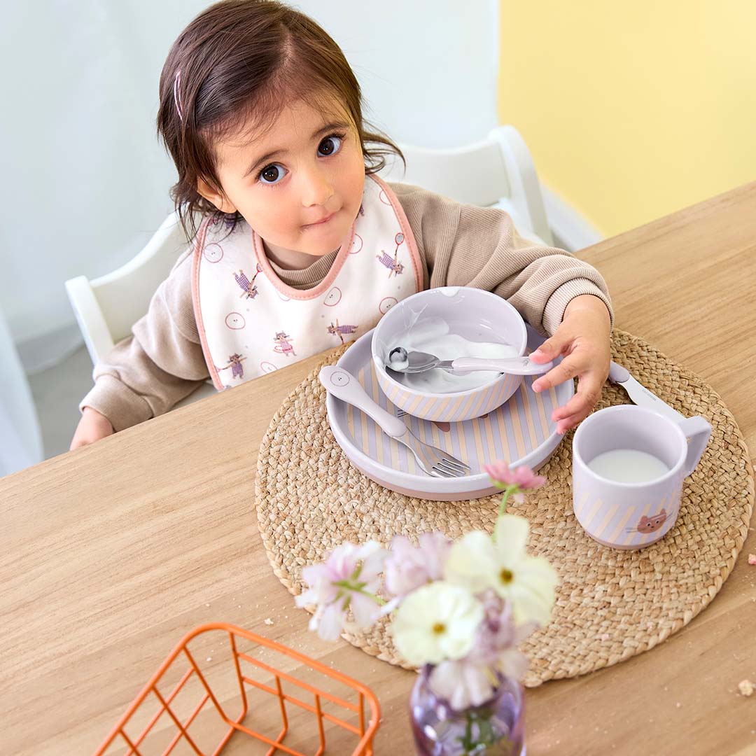 Een peuter zit aan tafel met een Lässig babyvoedingsset met gestreepte borden en een kattenmotief.