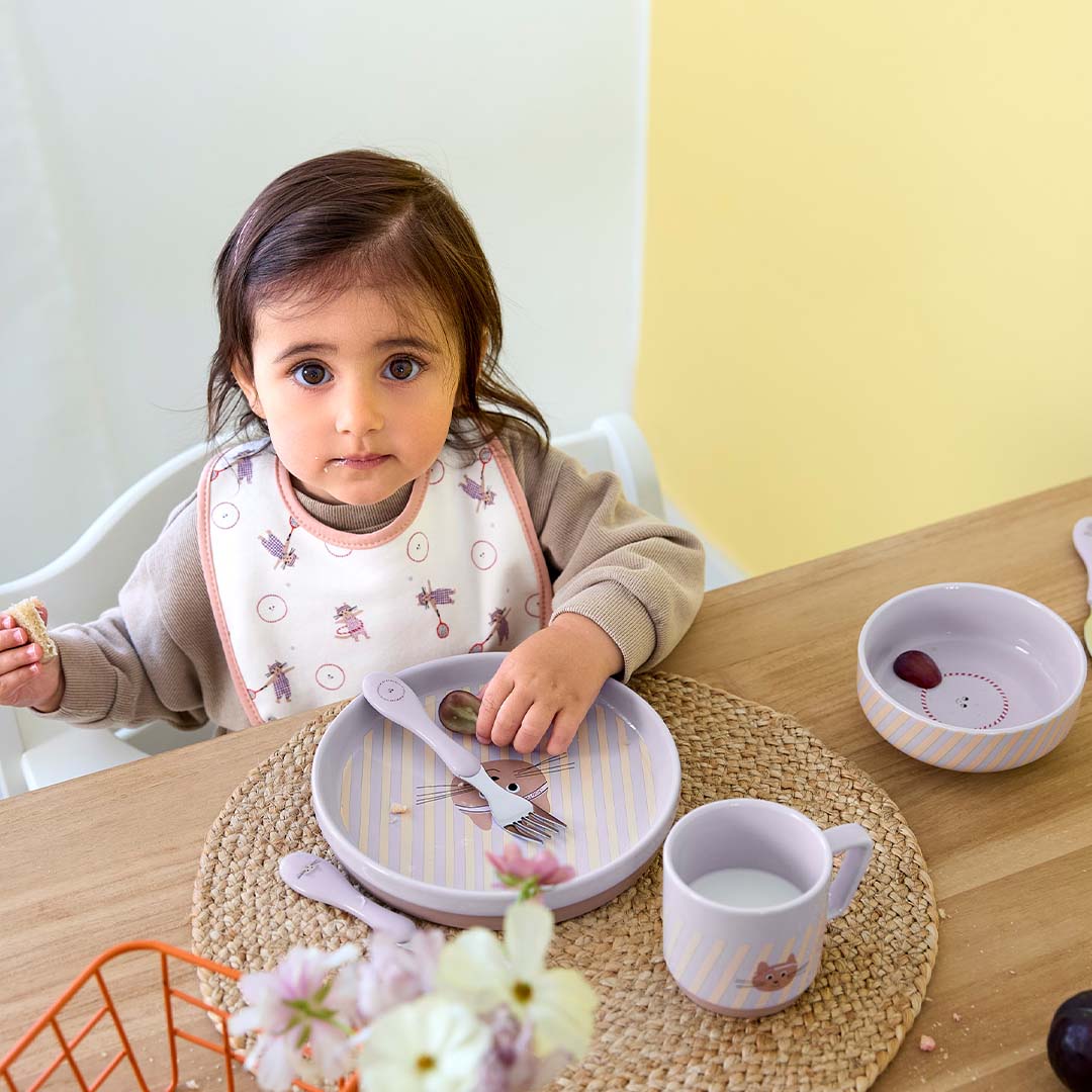 Een peuter zit aan tafel met een Lässig babyvoedingsset met gestreepte borden en een kattenmotief, en houdt een stukje brood vast.