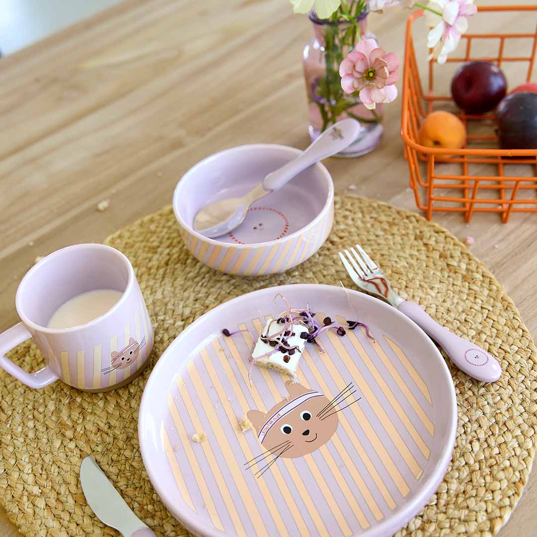 Een Lässig babyvoedingsset met een gestreept bord met kattengezicht, een gestreepte kom, een gestreepte mok en roze kinderbestek op een placemat.