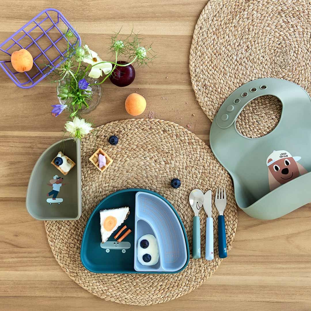 LÄSSIG kinderbestekset met bijpassende borden en een slabbetje op een houten tafel, gedemonstreerd met eten.