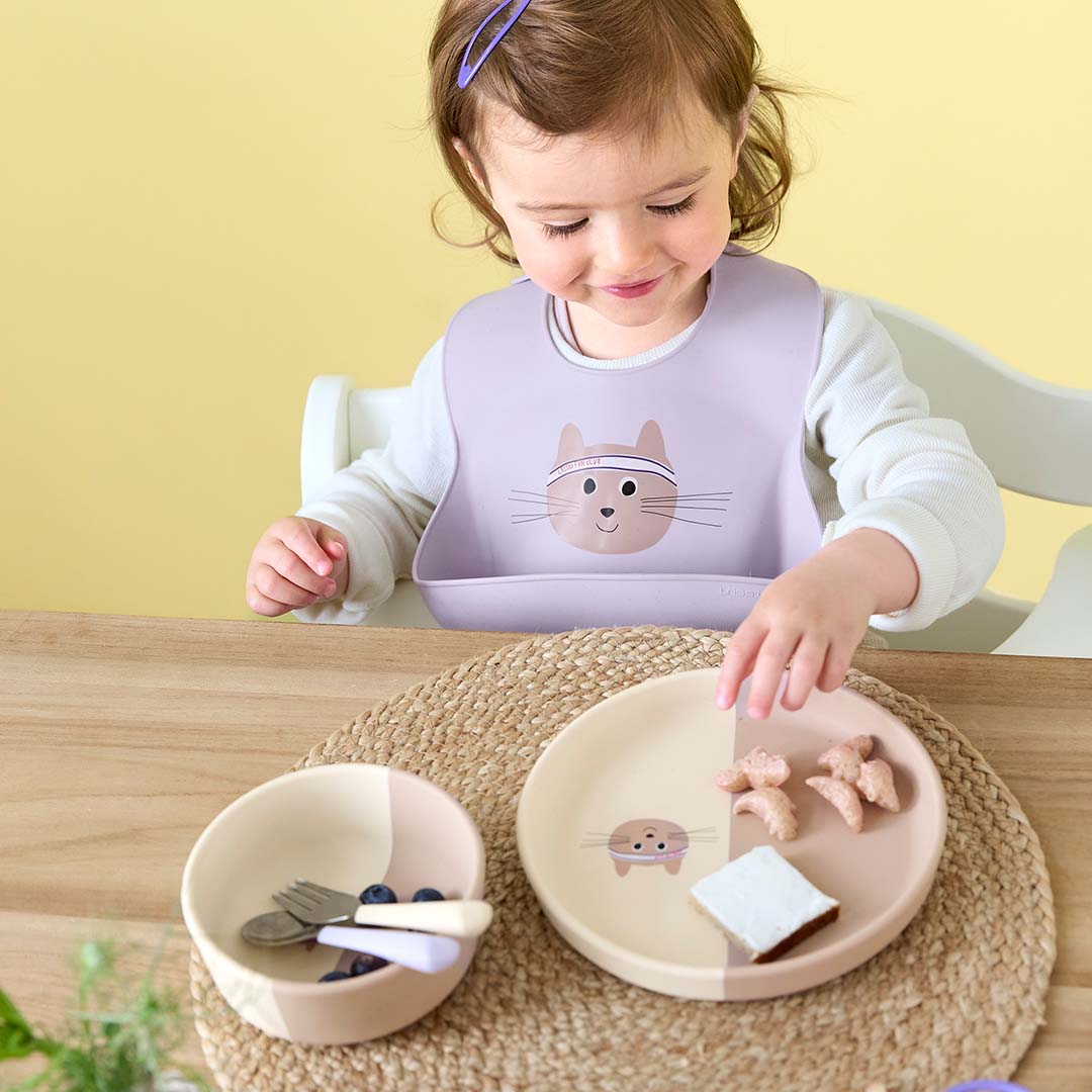 Een peuter met een paarse Lässig slab met kattengezicht eet aan tafel met bijpassend servies en kinderbestek.