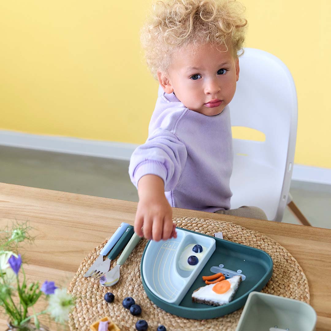Een kind aan tafel reikt naar een bord met eten, naast het lässig kinderbestek met groene handvatten.