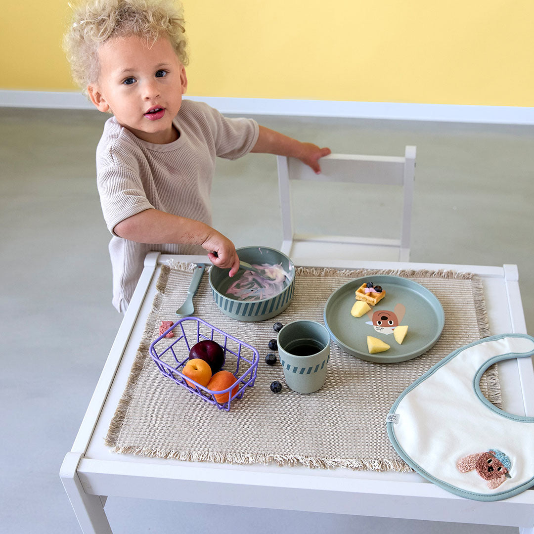 Een peuter staat naast een tafel met de groene lässig babyserviesset met hondenfiguur.
