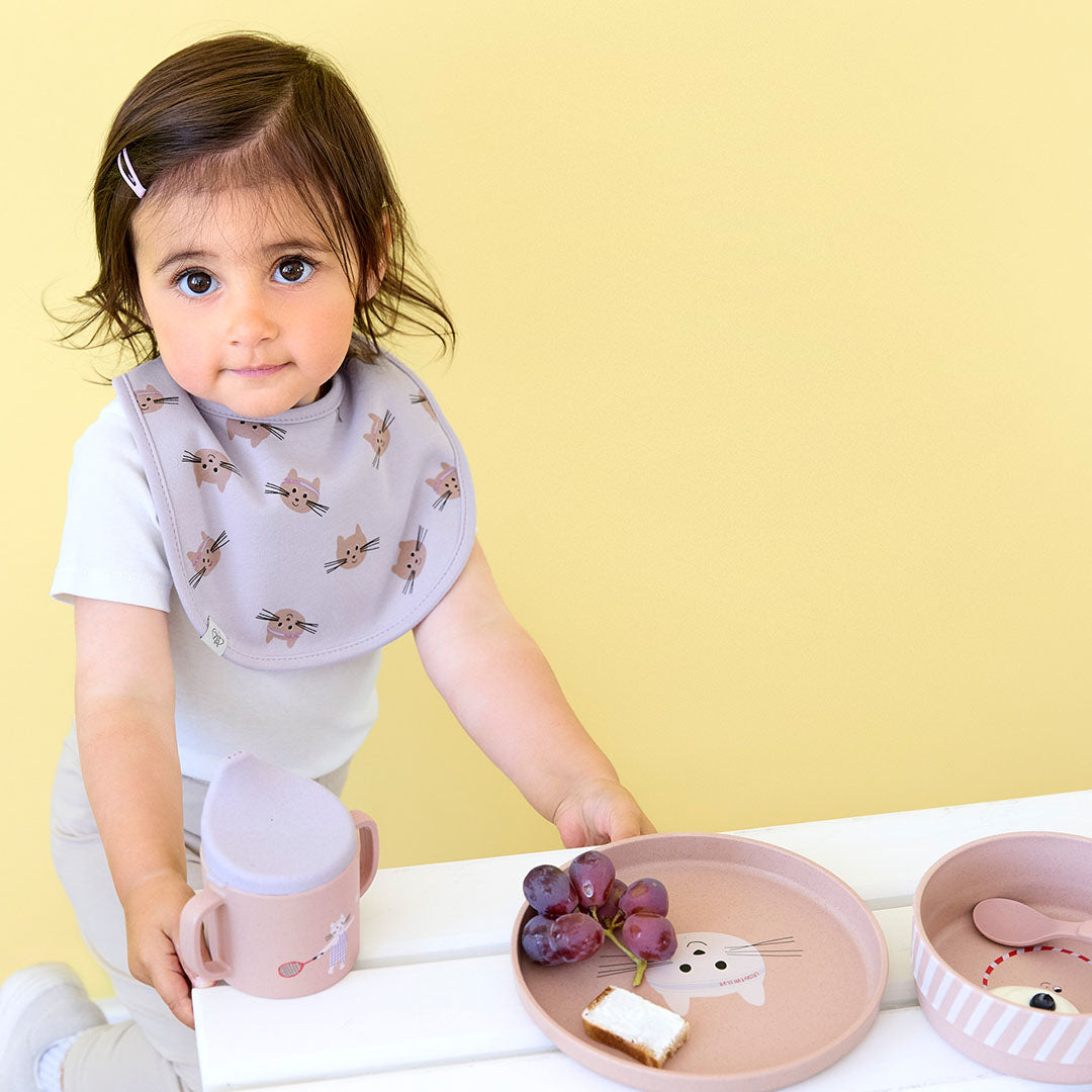 Een baby met een Lässig slabbetje met kattenprint staat naast een tafel met de roze serviesset en een bijpassende beker.
