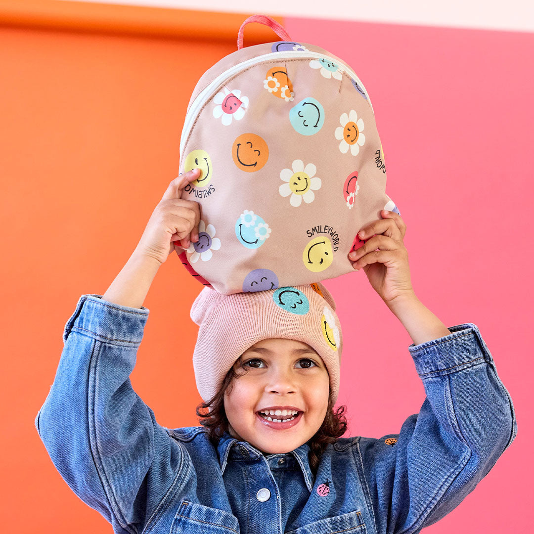 Een lachend meisje met een muts balanceert de Lässig SmileyWorld peuterrugzak op haar hoofd.