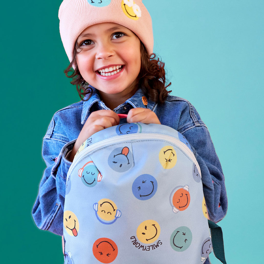Een lachend kind met een roze muts houdt de blauwe peuterrugzak met smileyworld-print vast.
