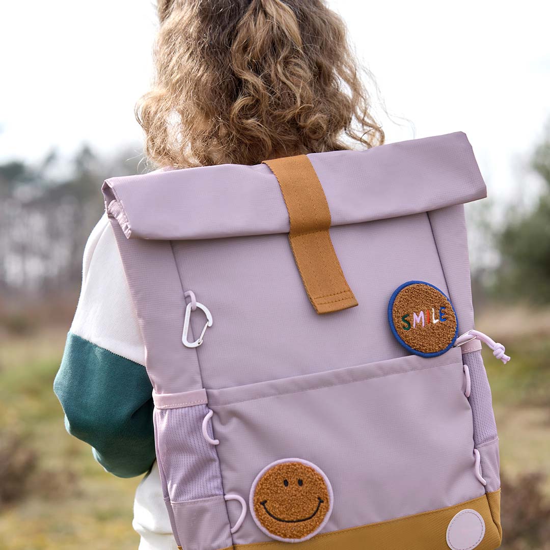 Kind draagt de lässig paarse schoolrugzak met smiley patch op de rug in een veld.