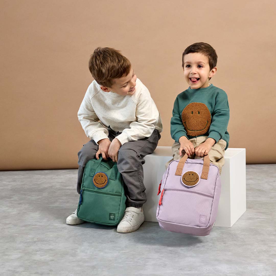 Twee lachende kinderen zitten met elk een Lässig kleuterrugzak, één groene en één paarse, beide met een smiley patch.