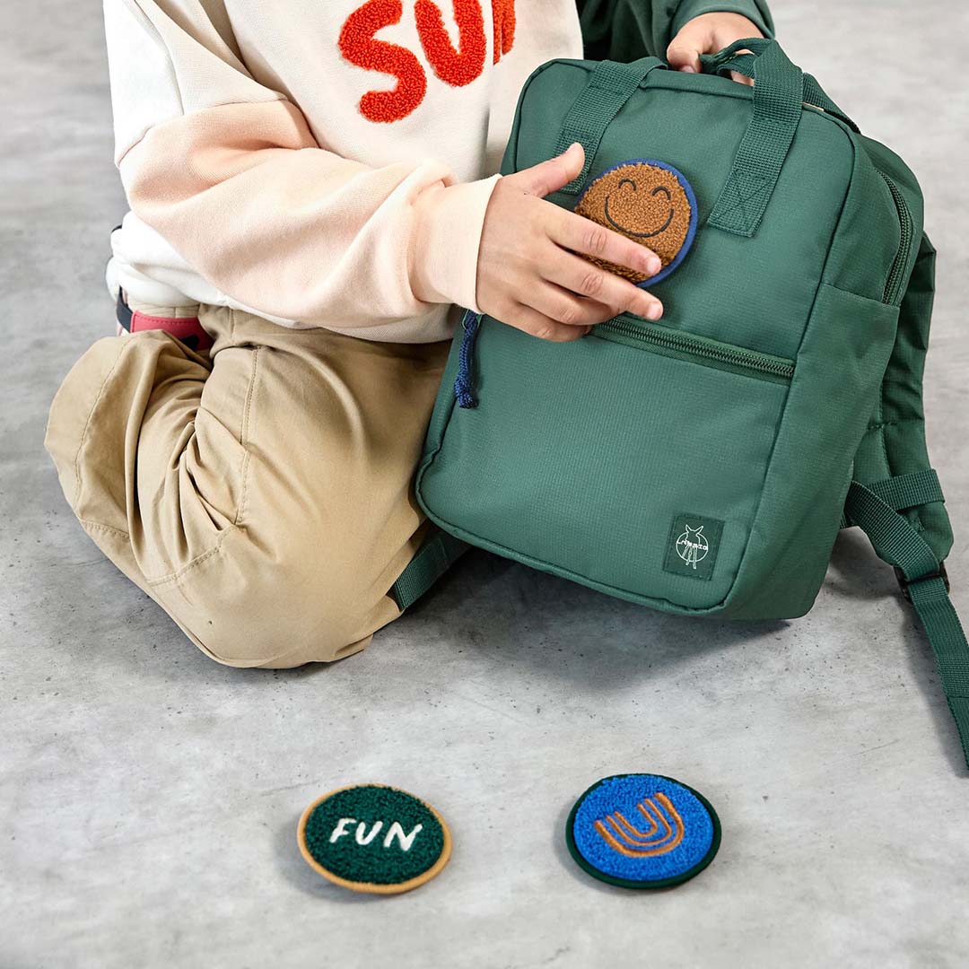 Een kind houdt de donkergroene Lässig kleuterrugzak vast, met een smiley patch en twee andere patches op de grond.