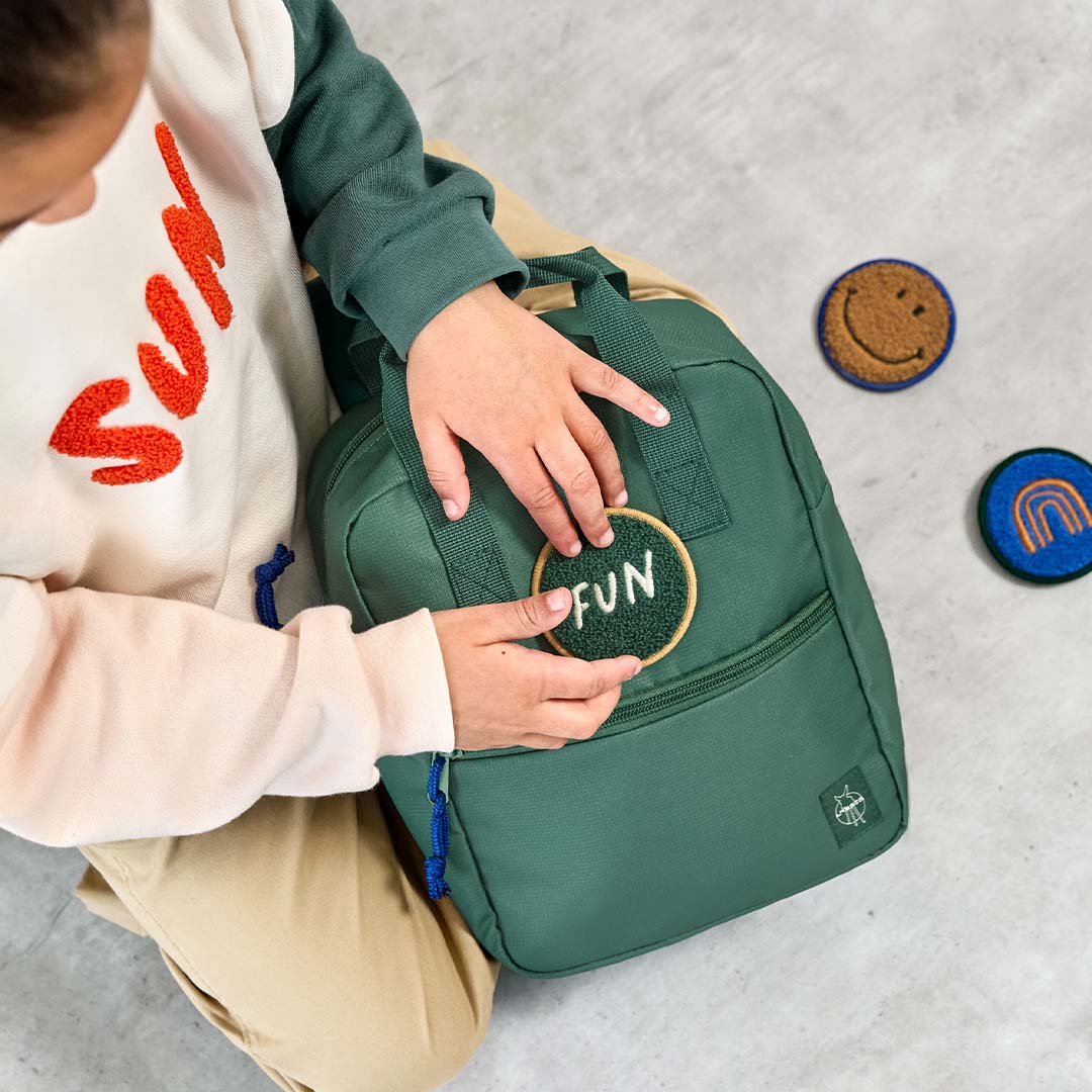 Kinderhanden plaatsen een 'FUN' patch op de donkergroene Lässig kleuterrugzak, met andere patches ernaast.