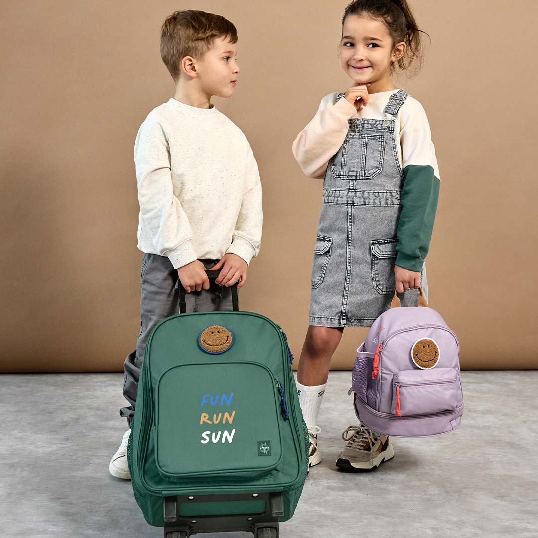 Een jongen met de lässig groene kinderkoffer en een meisje met een paarse kinderrugzak, beide met smiley patch.
