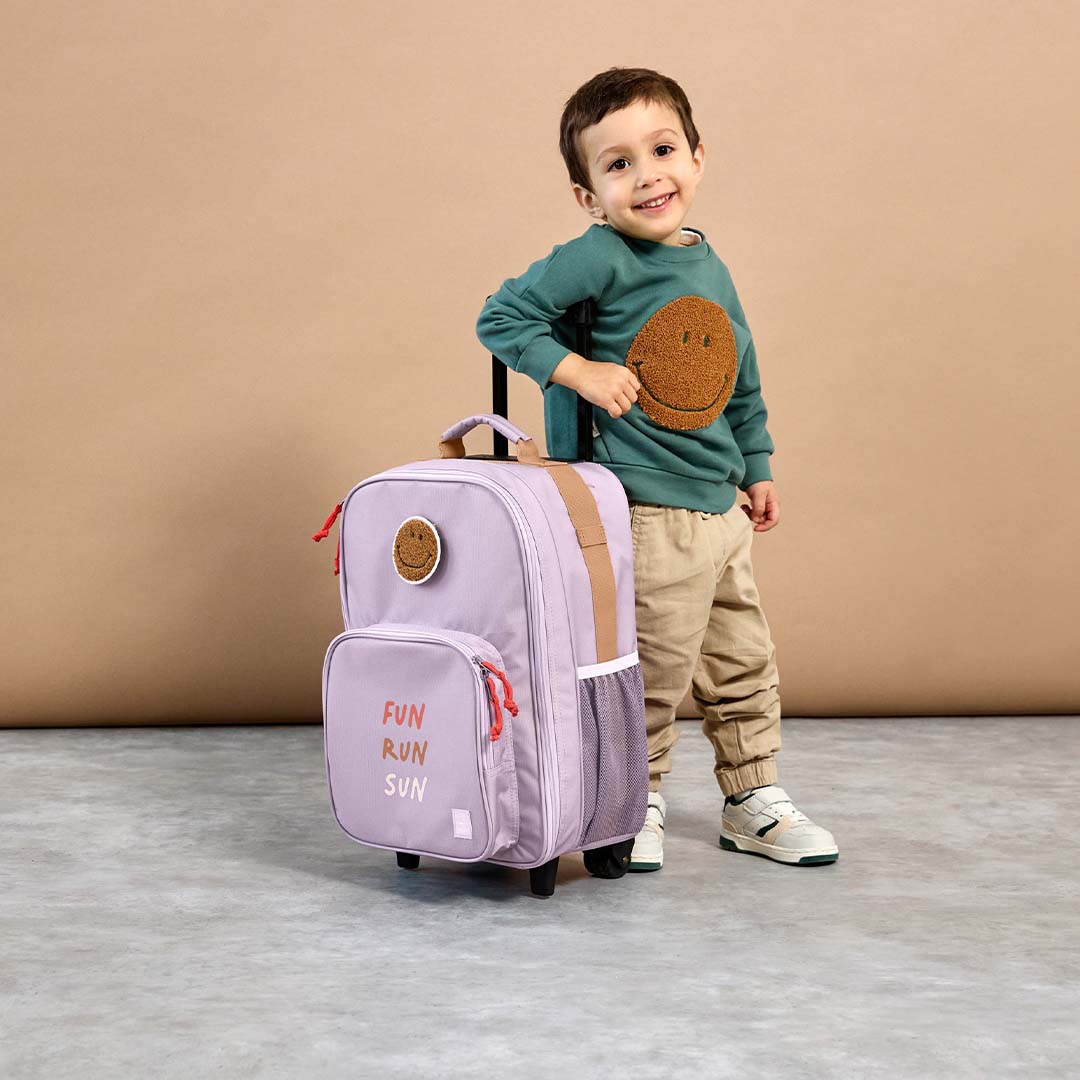 Een lachende jongen trekt de lässig kinderkoffer in lila met smiley patch.
