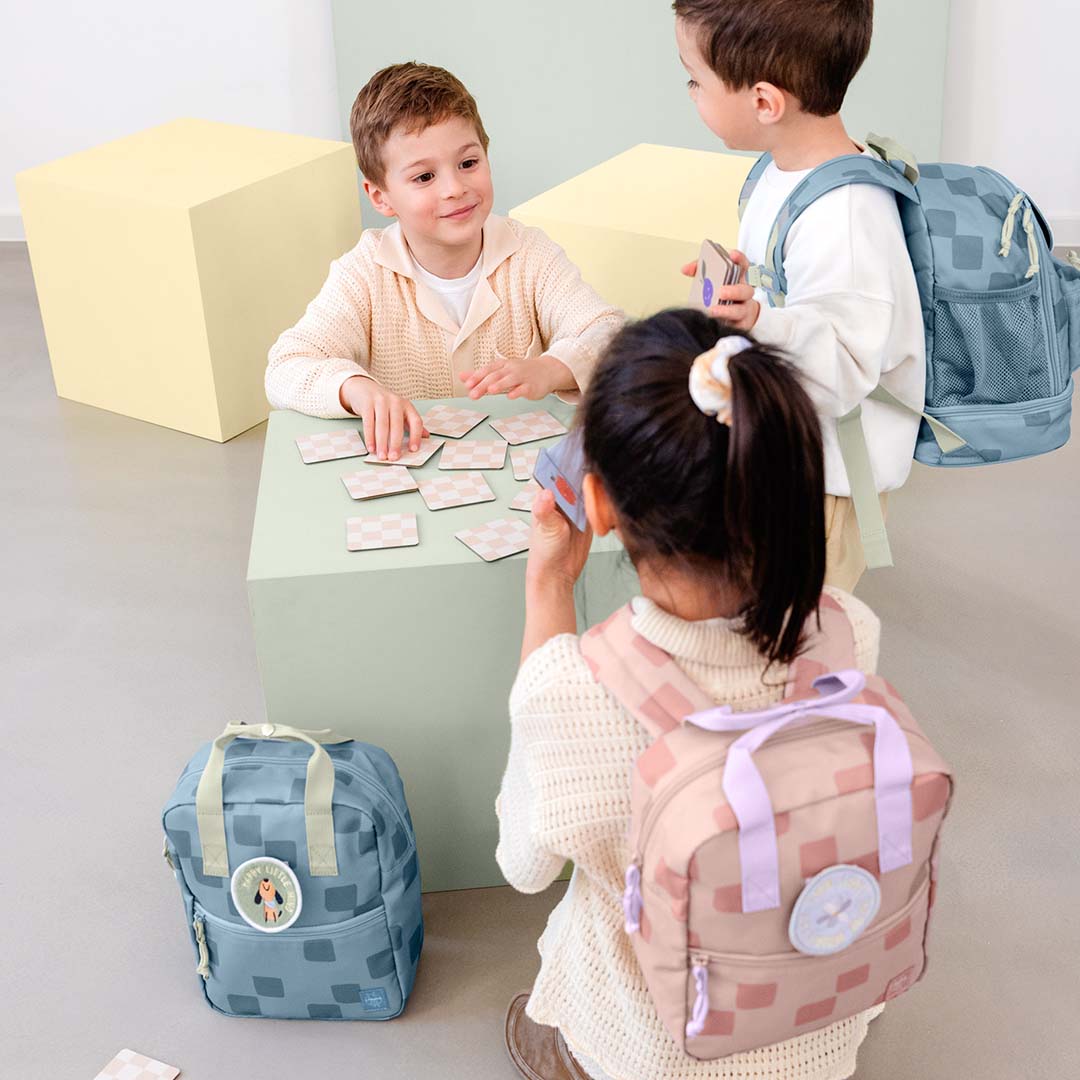 Twee kinderen spelen een spel, met een roze en een blauwe Lässig kleuterrugzak met geruit patroon op de grond.