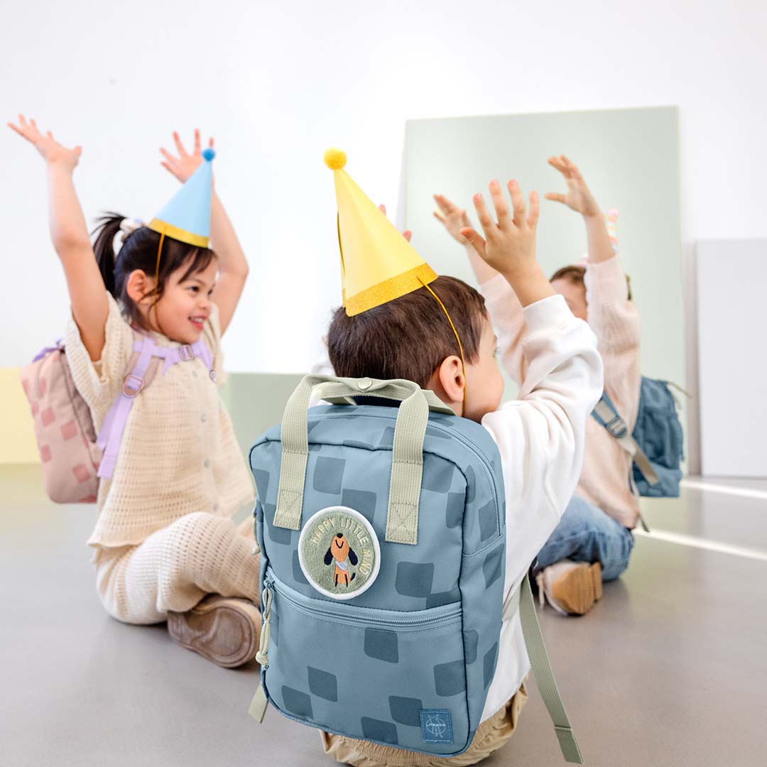 Twee kinderen met feestmutsen en rugzakken, waaronder de blauwe Lässig kleuterrugzak, spelen op de grond.