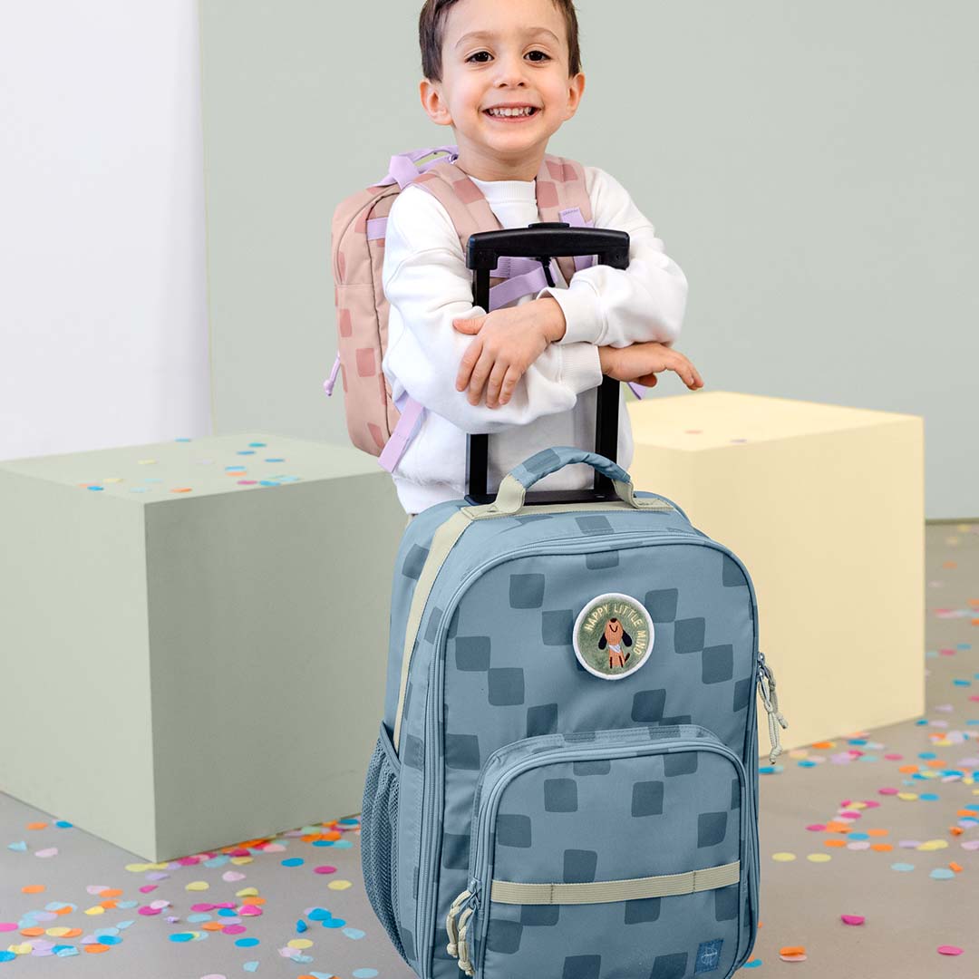 Een lachend kind staat achter de Lässig kinderkoffer met blauw geometrisch patroon.