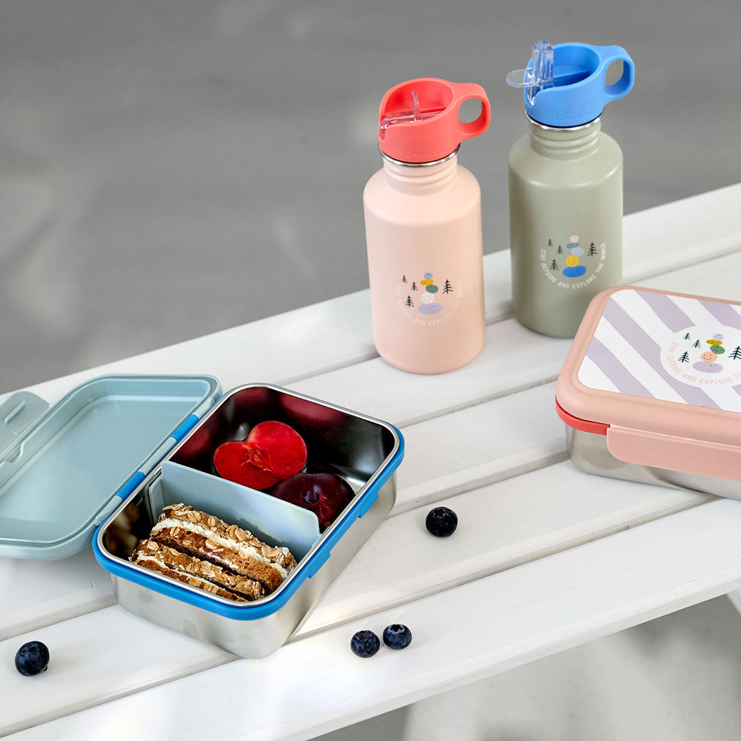 Twee Lässig kinder drinkflessen en twee bijpassende lunchboxen met fruit en koekjes op een witte tafel.