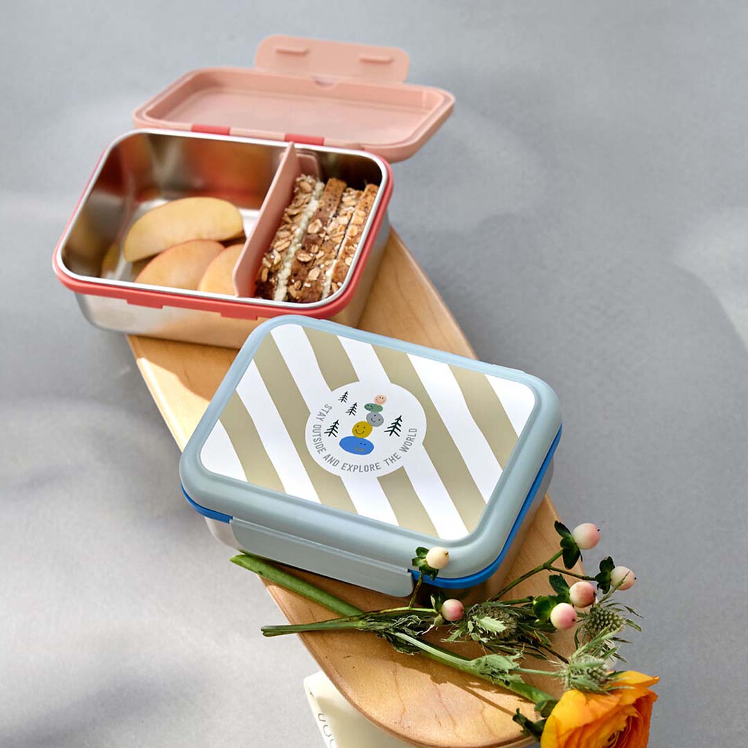 Twee Lässig rvs kinderbroodtrommels op een houten plank, één open met appel en brood, de andere gesloten met blauwe deksel.