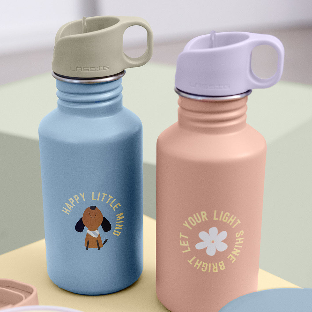 Twee Lässig kinderdrinkflessen van roestvrij staal, één roze met bloem en tekst, en één blauw met een hond en tekst.