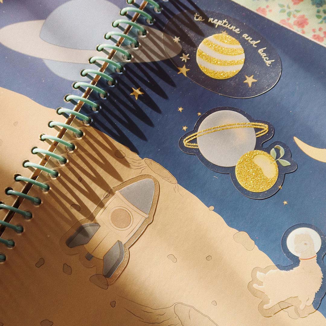 Detailfoto van een open Konges Sløjd stickerboek met ruimte-thema stickers van planeten, sterren en een raket op een donkerblauwe achtergrond.