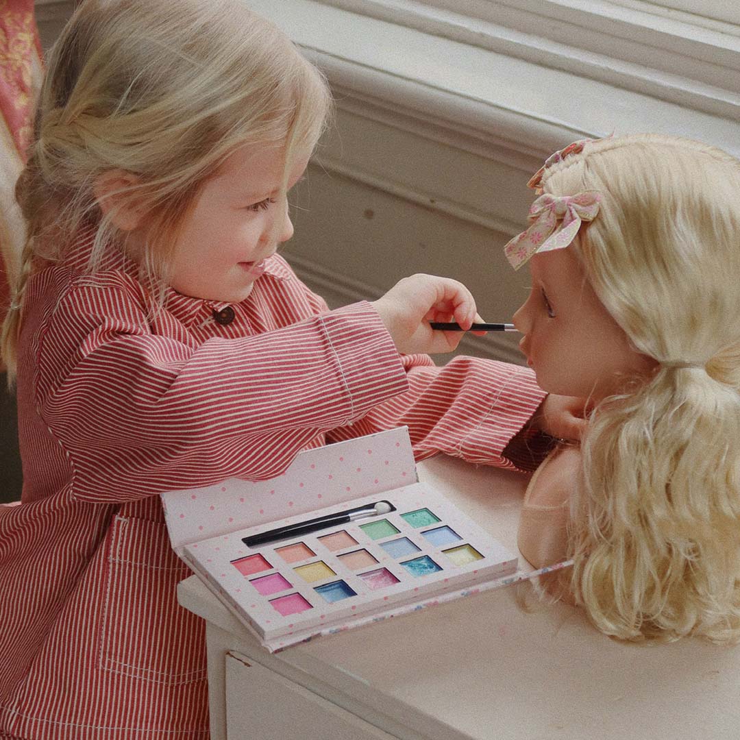 Een jong kind schminkt de Konges Sløjd make-up pop Lili met een kwastje en een make-up palet.