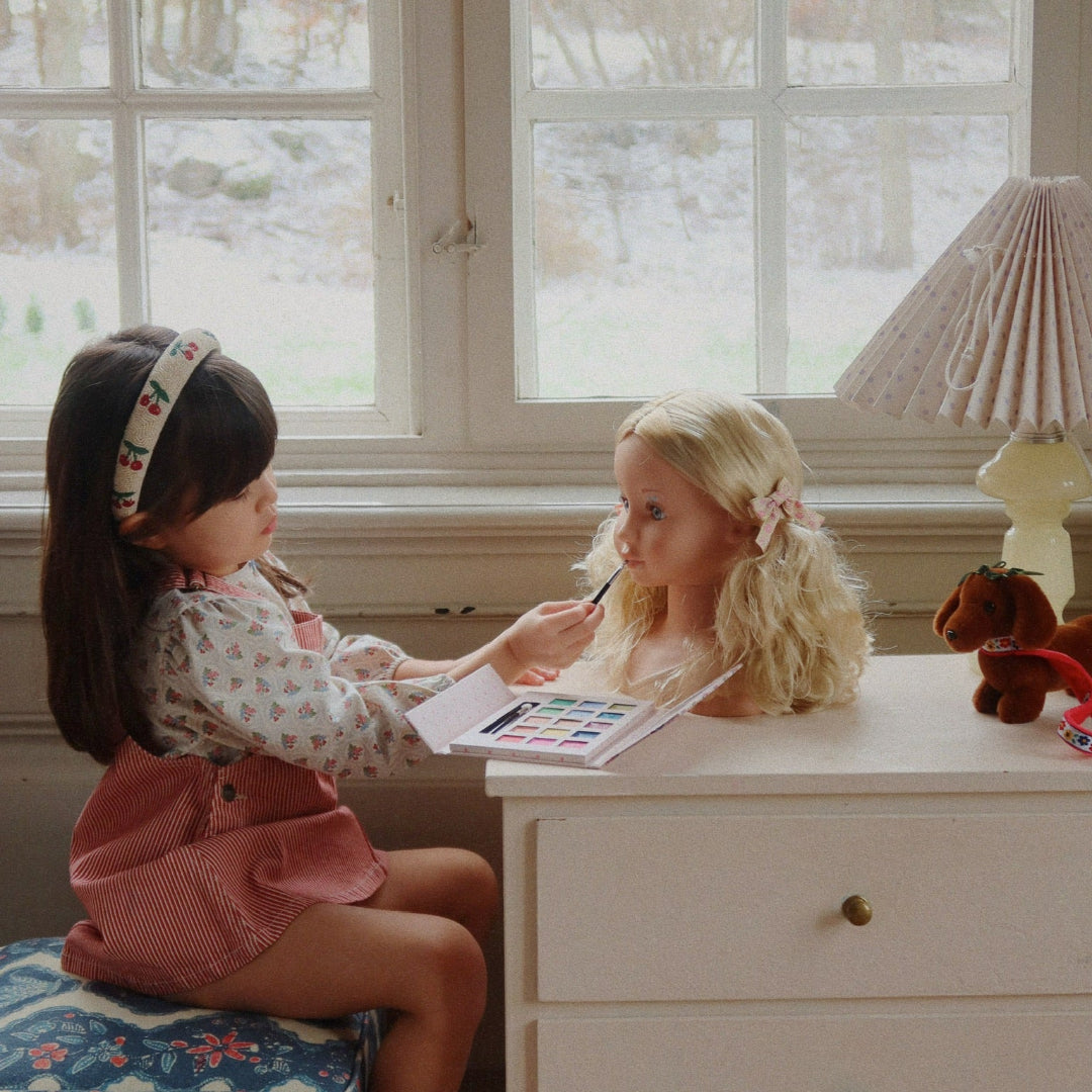 Een kind zit aan een tafel en schminkt de Konges Sløjd make-up pop Lili met een kwastje.