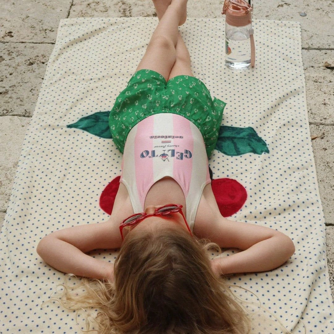 Een kind ligt op het Konges Sløjd strandlaken met kersenprint en stippen, naast een waterfles.