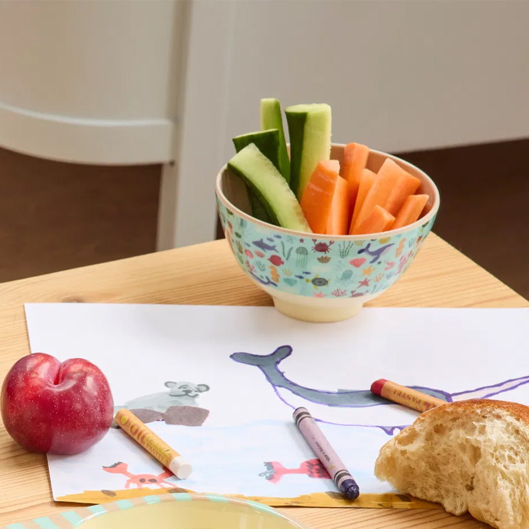 Kinderkom met zeedierenpatroon gevuld met wortel- en komkommerstaafjes op een houten tafel.
