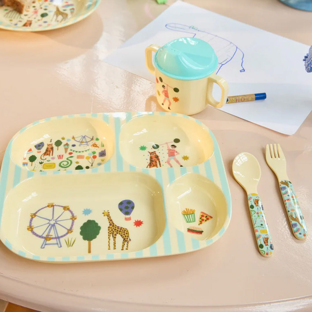 Het Rice melamine kinderservies met een verdeeld bord, beker, vork en lepel met kermis-thema op een roze tafel.
