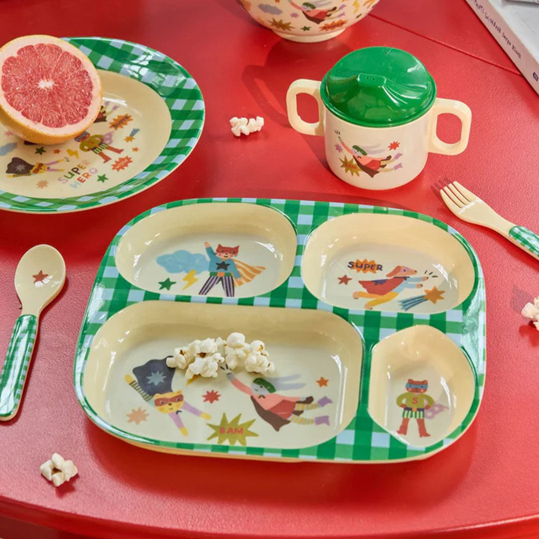 Rice kinderservies met superheldenfiguren en groen geruit patroon, gedemonstreerd op een rode tafel met popcorn en fruit.