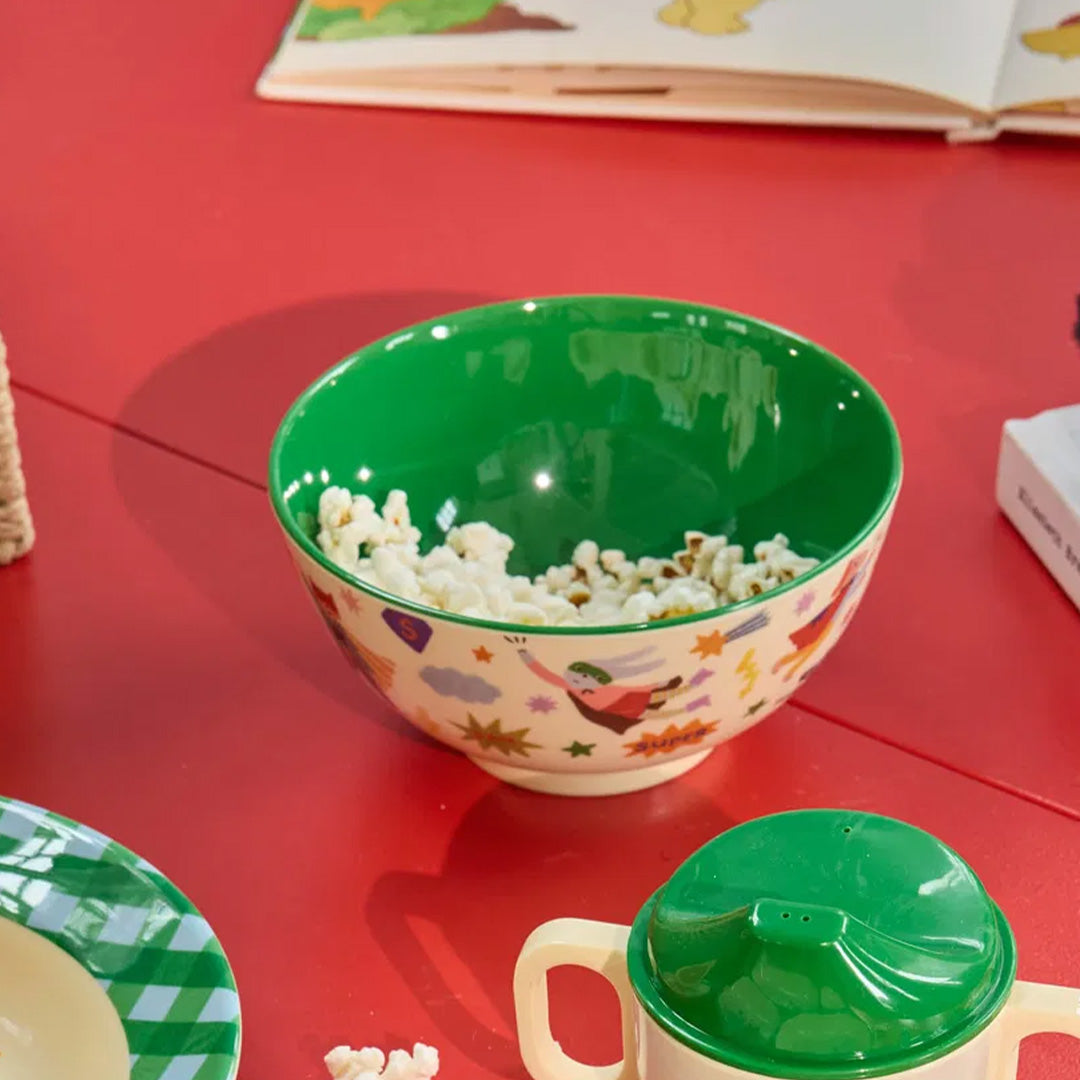 Kinderkom met superheldenprint gevuld met popcorn op een rode tafel.