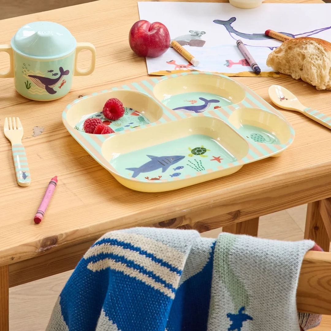 Het rice kinderservies met oceaanprint, bestaande uit een verdeeld bord, beker, vork en lepel, op een houten tafel.
