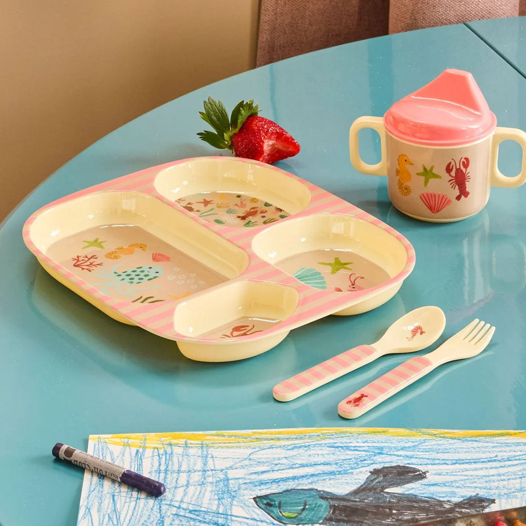 Het Rice kinderservies met onderwaterdieren en roze strepen, bestaande uit een verdeeld bord, drinkbeker en bestek, op een blauwe tafel.