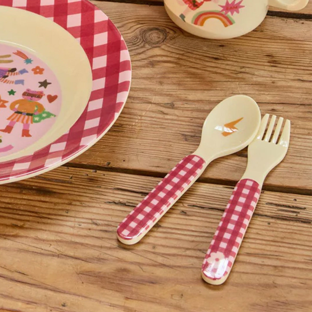 Kinderbestekset met rood-wit geruite handvatten, liggend op een houten tafel naast een bijpassend bord met hero-figuren.