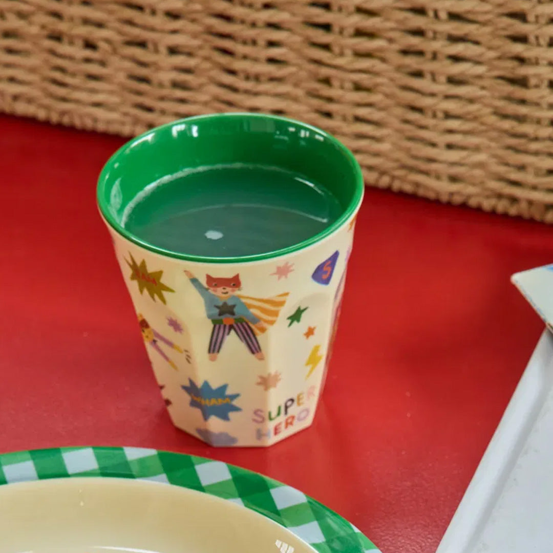 Kinderbeker met superheldenprint gevuld met groene vloeistof op een rode tafel.
