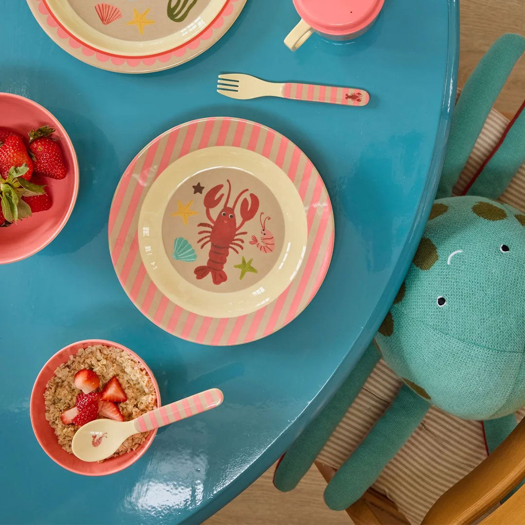 Rice kinderbestek met zeedieren op een blauwe tafel, naast een bord met eten en een knuffel octopus.