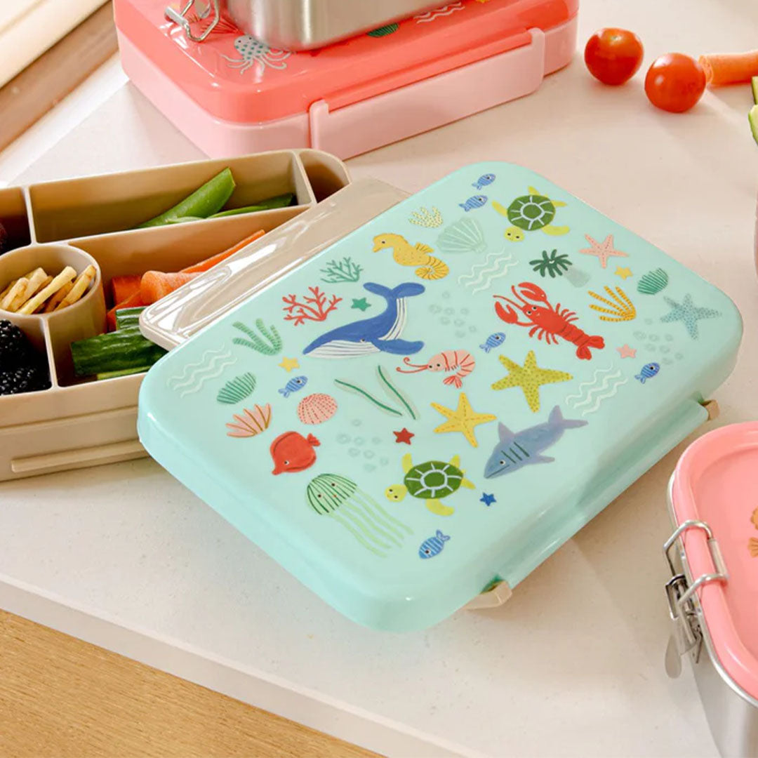 Een open bento broodtrommel met zeedieren op de deksel, gevuld met groenten en fruit op een aanrecht.
