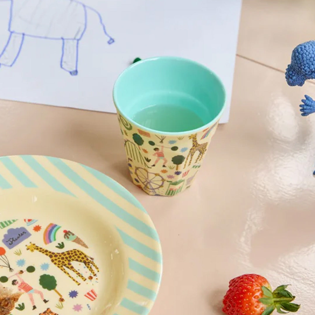 Een rice kinderbeker met fun fair print, gevuld met water, staat op een tafel naast een bord en een aardbei.