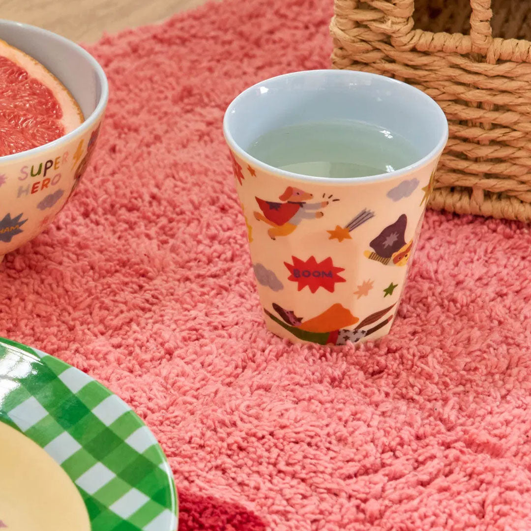 Kinderbeker met superheldenprint, gevuld met water, op een roze kleed naast een kom en bord.